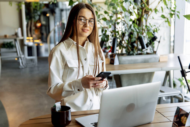 Charming female freelancer in trendy clothing working on laptop during morning breakfast at cozy cafe. Pretty young girls with brown hair smiling and posing on camera. - Fotografie, Obrázek