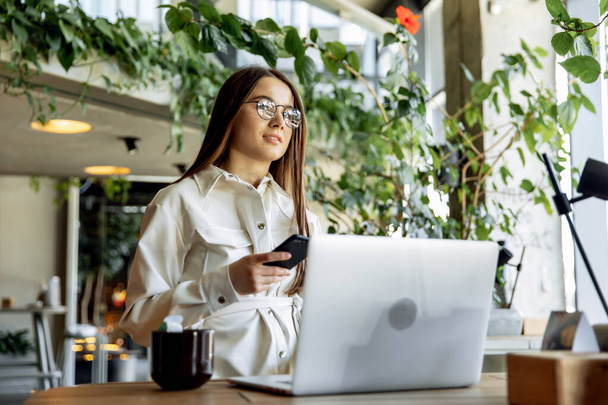 Charming female freelancer in trendy clothing working on laptop during morning breakfast at cozy cafe. Pretty young girls with brown hair smiling and posing on camera. - Foto, imagen