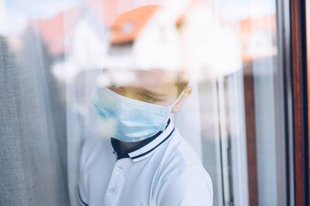 Niño triste con máscara médica mirando por la ventana. Cuarentena
 - Foto, imagen