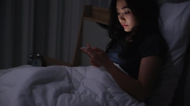 アジアの若い女性がベッドの上に寝そべって夜の時間帯にスマートフォンを再生します。携帯電話で社会を読む。低光で携帯電話を使用すると、目に健康への影響があります。不眠症の概念 - 映像、動画