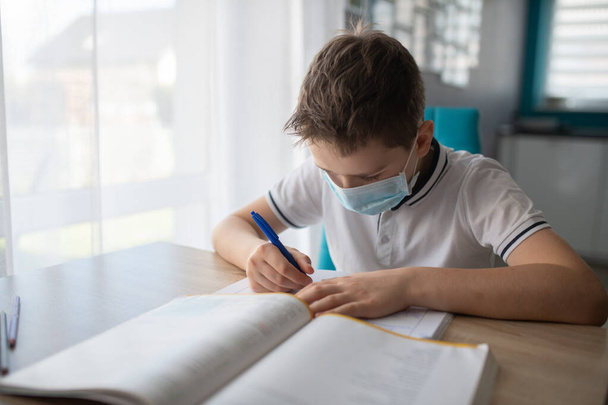 Παιδί με προστατευτική ιατρική μάσκα κάνει τα μαθήματά του. Το σχολείο έκλεισε κατά τη διάρκεια του Coronavirus - Φωτογραφία, εικόνα