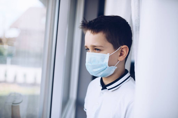 Ένα θλιμμένο παιδί με ιατρική μάσκα κοιτάζει από το παράθυρο. Καραντίνα - Φωτογραφία, εικόνα