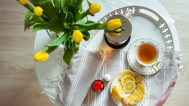 Plate of tea, lemon and ginger on bed table with yellow tulips becouse of corona virus - Foto, imagen
