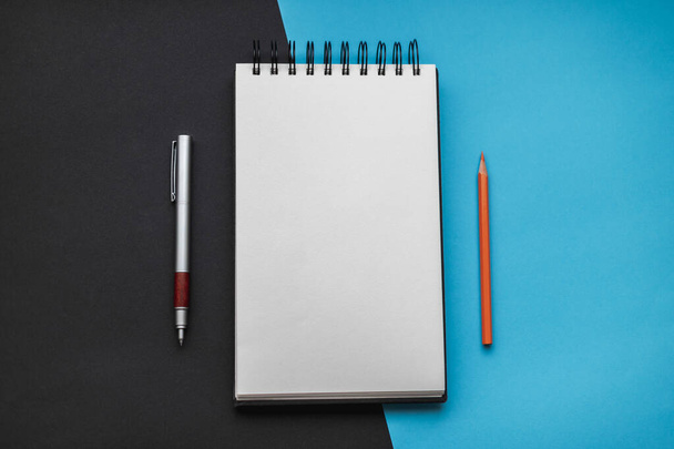 Open notebook with rings and a blank sheet in the middle, an orange pencil pointing up on the right and a metal pen pointing down on the left, all together with a black and blue background cut diagonally. - Fotoğraf, Görsel