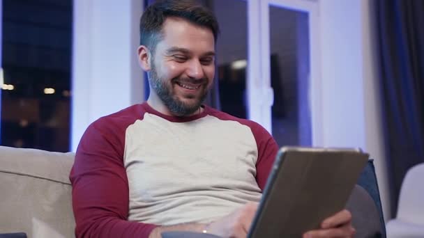 Front view of smiling satisfied young bearded guy which sitting on the couch and using tablet pc for review funny videos - Metraje, vídeo