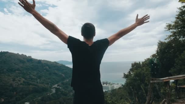 Νεαρός ταξιδιώτης σηκώνει τα χέρια του ψηλά στην κορυφή του βουνού πάνω από το όμορφο τοπίο - Πλάνα, βίντεο