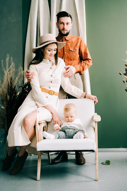Bej elbiseli ve şapkalı güzel bir anne, şık giyimli oğlunu kollarında tutuyor. - Fotoğraf, Görsel