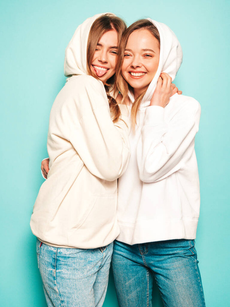 Zwei junge schöne blonde lächelnde Hipster-Mädchen in trendigen Sommer-Kapuzenpullovern. Sexy unbeschwerte Frauen posieren in der Nähe der blauen Wand. Modische und positive Modelle, die Spaß haben. - Foto, Bild