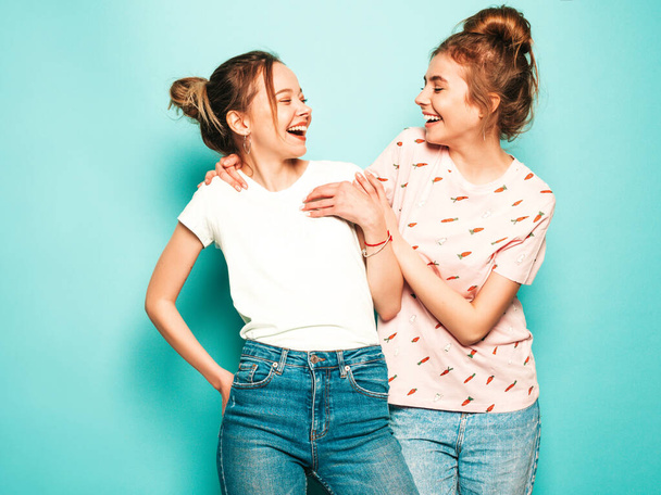 Две молодые красивые блондинки улыбающиеся хипстеры в модных летних хипстерских джинсах. Сексуальные беззаботные женщины позируют у голубой стены. Традиционные и позитивные модели развлечения - Фото, изображение