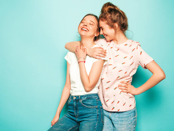 Twee jonge mooie blonde lachende hipster meisjes in trendy hipster jeans kleding. Sexy zorgeloze vrouwen poseren naast de blauwe muur. Trendy en positieve modellen hebben plezier - Foto, afbeelding