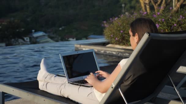girl texts on laptop resting at hotel pool against bush - Felvétel, videó