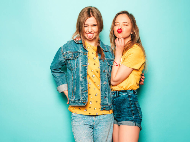 Two young beautiful blond smiling hipster girls in trendy summer hipster jeans clothes. Sexy carefree women posing near blue wall. Trendy and positive models having fun.They show tongues - Foto, Imagem