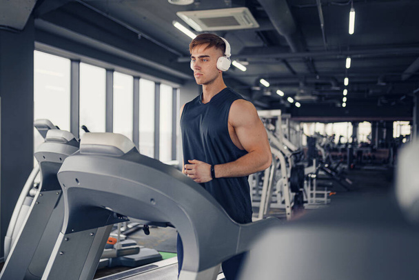 Concentrated fit man listening to music in headphones while running on treadmill in modern gym with panoramic windows. - Photo, Image