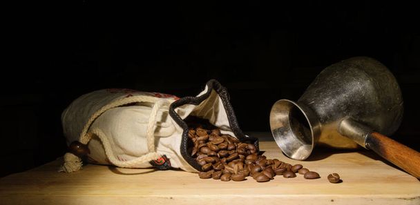 Σε μια υφασμάτινη τσάντα υπάρχουν κόκκοι καφέ. Οι κόκκοι είναι διάσπαρτοι σε μια ξύλινη σανίδα. Κοντινό πλάνο. Μαύρο φόντο. Εδώ κοντά είναι μια μεταλλική καφετιέρα. Concept - διάλειμμα για καφέ, αγάπη για τον καφέ - Φωτογραφία, εικόνα