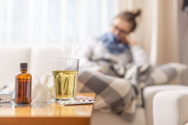 Young ill woman lying under blanket in a bed with seasonal illnes drinking hot tea with honey and lemon. She takes pills and cough sirup to push the disease. - Фото, зображення