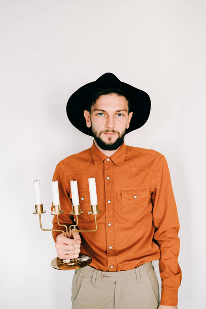 молодой стильный парень в оранжевой рубашке и шляпе с канделябрами в руках
 - Фото, изображение
