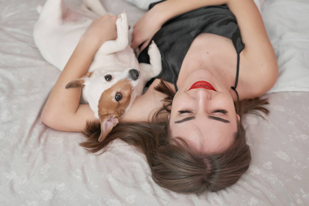 Girl and dog in bed. Beautiful woman in pajamas sits on bed in bedroom with dog Jack Russell Terrier, enjoys morning weekend, has good relationship with pet. Man best friend dog. Cozy morning. - Фото, зображення