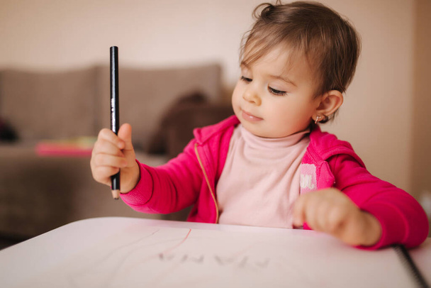 Glückliche kleine Mädchen sitzen am Tisch und winken etwas. Kleines Mädchen zeichnet zu Hause mit Bleistift auf weißem Papier - Foto, Bild