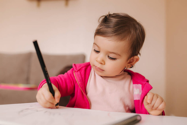 Gelukkige kleine meid zit aan tafel en wthire iets. Klein meisje gebruik potlood voor het tekenen op wit papier thuis - Foto, afbeelding