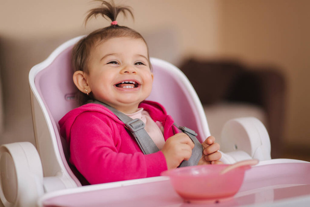 Маленька мила дівчинка їсть у високому кріслі. Портрет маленької усміхненої дитини вдома
 - Фото, зображення