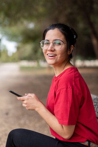 Mladá atraktivní žena s brýlemi sedí na lavičce a používá telefon v parku, s červenou košilí. Sevilla, Španělsko - Fotografie, Obrázek