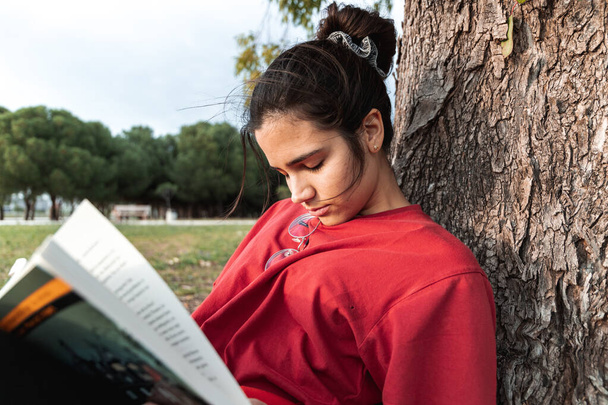 Giovane donna che dorme mentre legge un libro e si annoia. Indossa una camicia rossa e ha gli occhiali al collo. Dorme con la schiena su un albero alto
. - Foto, immagini
