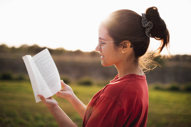 Mujer joven que tiene una buena lectura mientras sonríe y disfruta de la luz de una puesta de sol en un parque
. - Foto, imagen