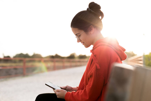 Eine junge Frau mit Brille in ihren Zwanzigern plaudert auf einer Parkbank mit ihrem Handy. Sie lächelt und trägt einen roten Pullover und einen Pferdeschwanz. Sie ist bei Sonnenuntergang mit Tageslicht beleuchtet - Foto, Bild
