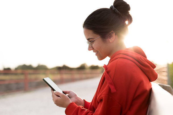 Young woman with glasses on her twenties chatting on her phone while sitting in a bench of a park. She is smiling and wearing a red sweater and a ponytail. She is backlit with daylight at sunset - Φωτογραφία, εικόνα