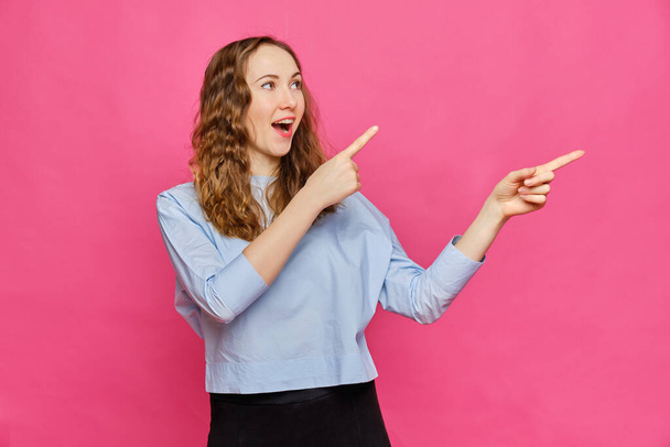 Stylish caucasian girl in a pale blue t-shirt laughs and shows fingers to the side on a pink background. Copy space. Close up. - Foto, Imagem