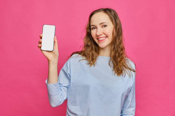 Stylish caucasian girl in a pale blue t-shirt shows smartphone on a pink background. Copy space. Close up. - Foto, Bild