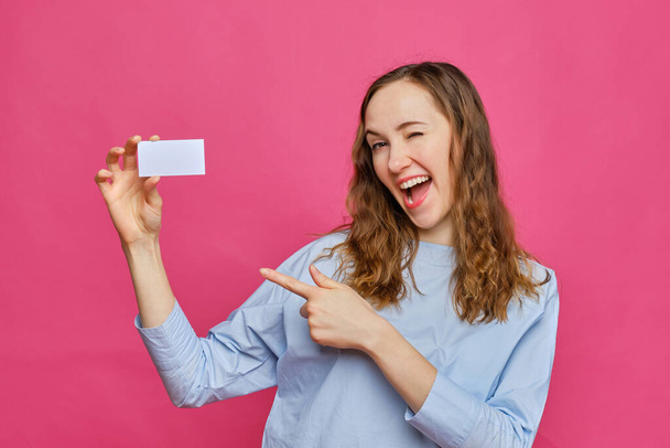 Stylish caucasian girl in a pale blue t-shirt hands holding a white card and points to it on a pink background. Copy space. Close up. - Photo, image