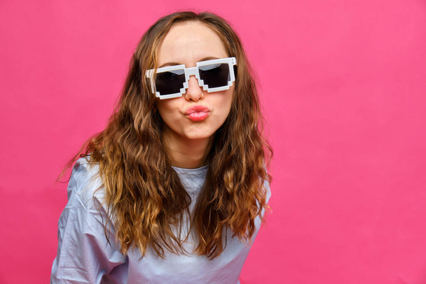 Stylish caucasian girl in a pale blue t-shirt and 8-bit glasses shows a kiss with lips on a pink background. Close up. - Photo, Image