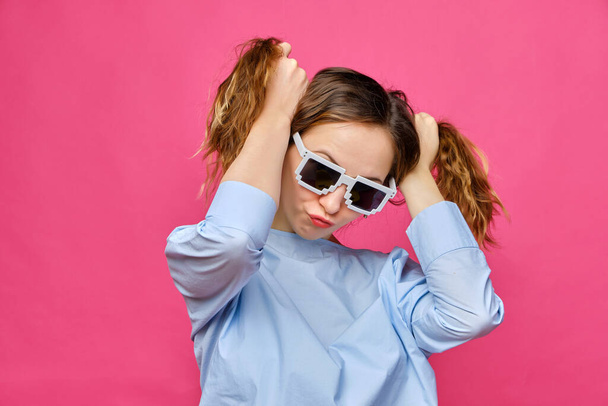 Stylish caucasian girl in a pale blue t-shirt and 8-bit glasses and makes two tails of hair on a pink background. Close up. - 写真・画像