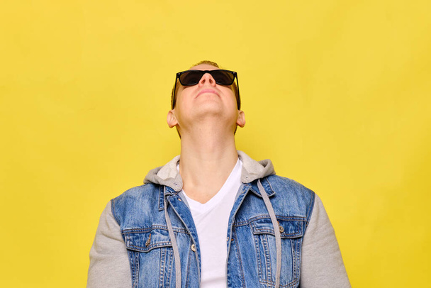 Stylish Caucasian man in a jeans and sunglasses on a yellow background. Copy space. Close up. Raised his head looking up. - Valokuva, kuva