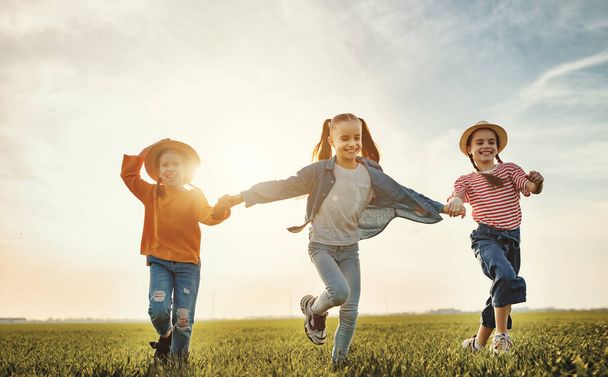 Веселі діти дівчата посміхаються і бігають на зеленій траві, розважаючись на лузі під час заходу сонця
 - Фото, зображення