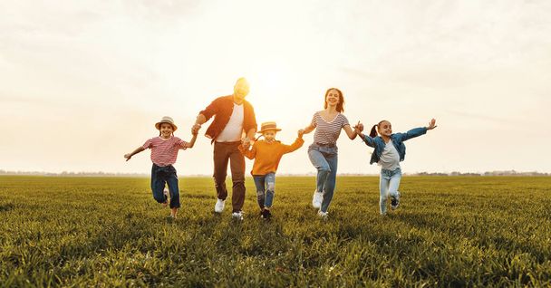 Weseli rodzice z dziećmi uśmiechniętymi i biegającymi po zielonej trawie podczas zabawy na łące podczas sunse - Zdjęcie, obraz