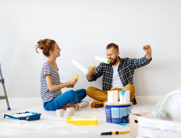 Cheerful couple young woman sitting crossed legged on floor and laughing while man singing into paint roller against blank white wall during renovatio - Foto, Bild