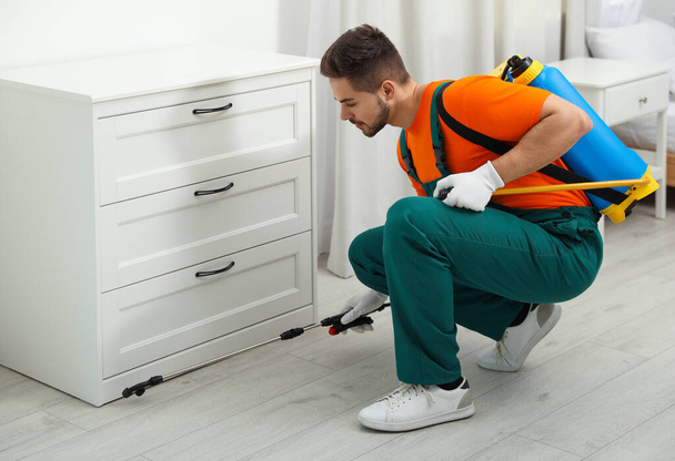 Pest control worker spraying insecticide near chest of drawers indoors - Foto, afbeelding