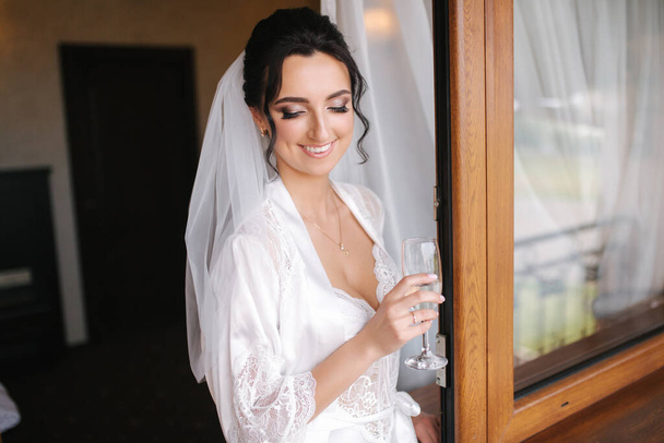 Portrait of gorgeous bride with glass of champagne at hotel in morning. Charming woman with great hairstyle and makeup - Photo, Image