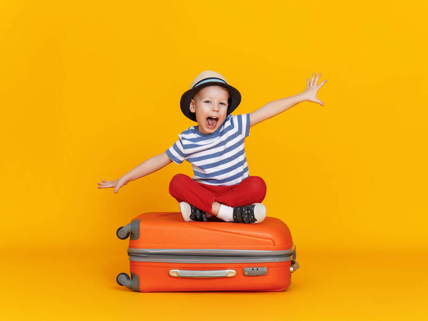 Ευτυχισμένο μικρό αγόρι με απλωμένα χέρια και ανοιχτό στόμα κάθεται σταυροπόδι στις αποσκευές και προσποιείται ότι πετάει εναντίον κίτρινο backgroun - Φωτογραφία, εικόνα