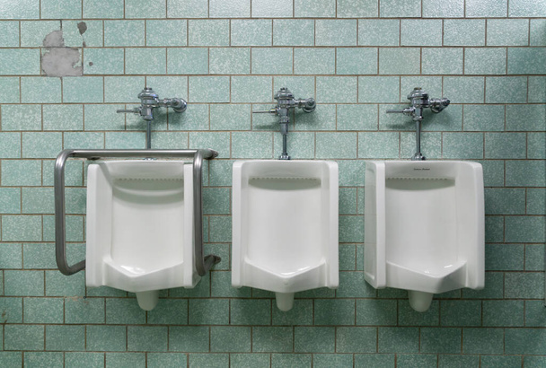 Horizontal interior image of three urinals mounted on a tile wall. One urinal is equipped for handicapped. - Photo, Image