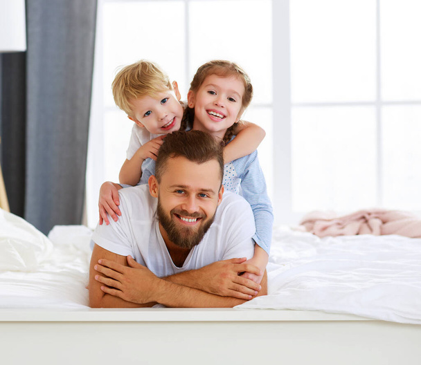 Оптимістичний бородатий чоловік тато з щасливими дітьми син і дочка на спині посміхаються і дивляться на камеру, лежачи на зручному ліжку в Морніні
 - Фото, зображення