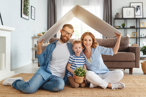 Vrolijke ouders met kind glimlachen en houden dak mockup over hoofden tijdens het zitten op de vloer in gezellige woonkamer tijdens relocatio - Foto, afbeelding