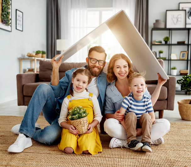 Cheerful parents with children smiling and keeping roof mockup over heads while sitting on floor in cozy living room during relocatio - Foto, immagini