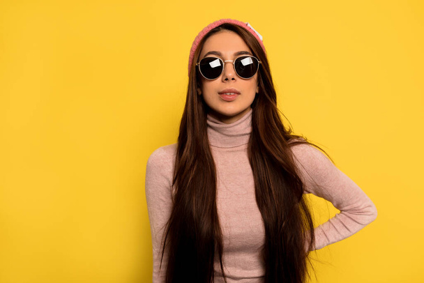 黄色の背景にピンクのキャップと黒のメガネを身に着けている長い暗い髪を持つ流行のスタイリッシュな女性の屋内スタジオショット  - 写真・画像