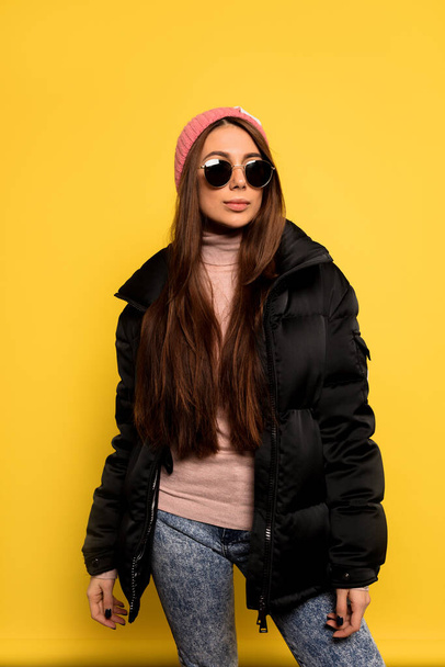 Fashion pretty carefree woman wearing a colorful pink cap and black jacket over yellow background. Appealing girl with long hair posing in studio - Foto, afbeelding