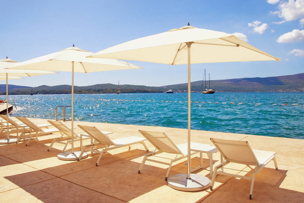 Férias na praia. Cadeiras de estar e guarda-sóis brancos contra o céu azul. Montenegro, Mar Adriático, Baía de Kotor, cidade de Tivat
  - Foto, Imagem