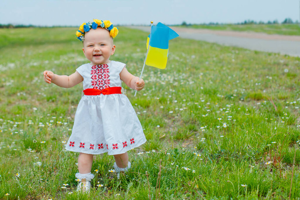 Pieni tyttö valkoisessa mekossa Ukrainan kirjonta, kirjonta, kävely pellolla koiranputkea ja kuljettaa Ukrainan lipun. Lapsi heiluttaa kelta-sinistä lippua. Onnellinen lapsi itsenäisyyspäivänä Ukrainan ja lipun päivä. - Valokuva, kuva