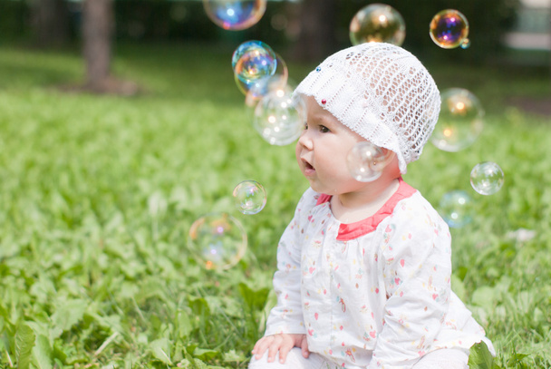 een klein meisje, zittend op groen gras in het park kijken naar zeep - Foto, afbeelding